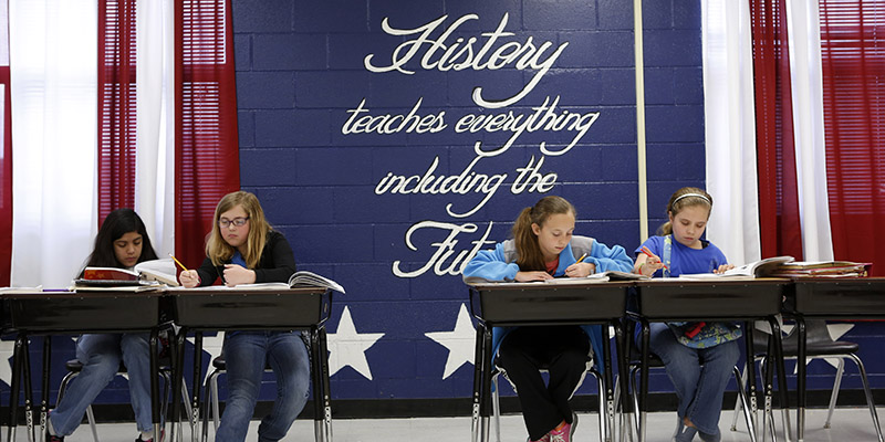 Four students reading at their desks. Behind them is a red, white and blue wall with the saying, History teaches everything, including the future.