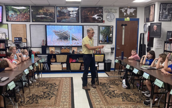 Donnie Wilkerson stands in the middle of his classroom talking to his students, who are seated at tables running along both sides of the walls of the classroom, which are covered in historical paintings and documents.