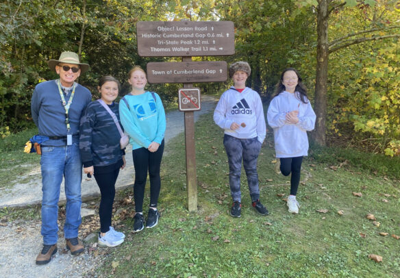 Donnie Wilkerson stands outside with four of his students at a trail marker in a forest. Trail marker reads: Object Lesson Road; Historic Cumberland Gap, 0.6 miles; Tri-State Peak, 1.2 miles; Thomas Walker Trail, 1.1 miles; Town of Cumberland Gap.