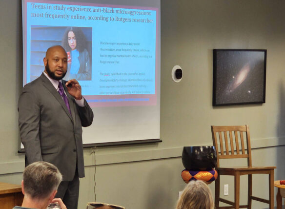 Kumar Rashad, dressed in a suit and tie, stands in front of a classroom talking to students. A screen behind him displays an article titled, "Teens in study experience anti-black microaggressions most frequently online, according to Rutgers researcher."