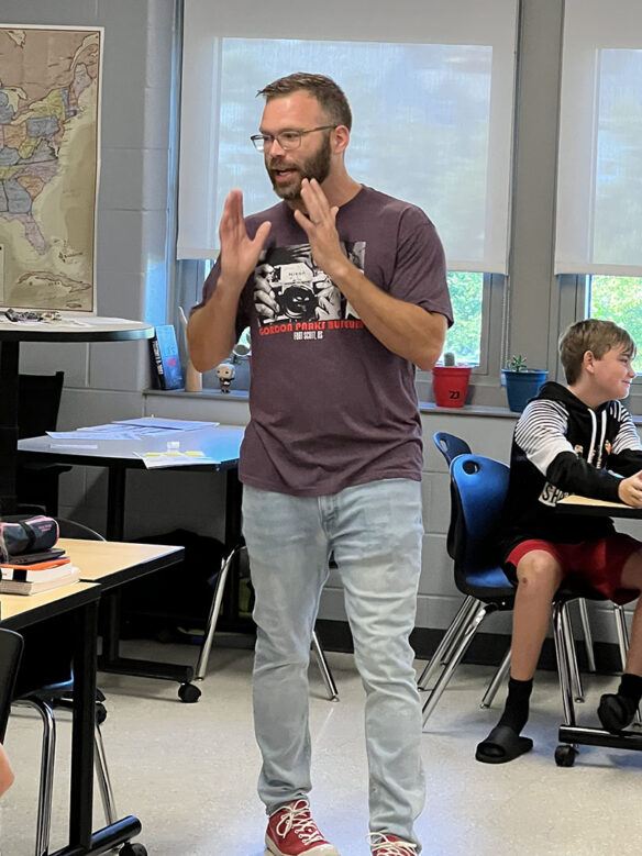 Kevin Dailey gestures with his hands up in the air while teacher in his classroom.