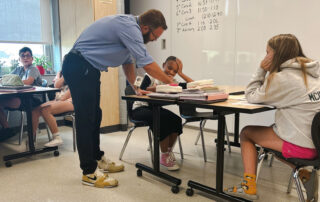 A picture of Kevin Dailey in his classroom, bending over a desk to help a young student.