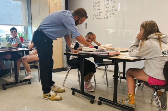 A picture of Kevin Dailey in his classroom, bending over a desk to help a young student.