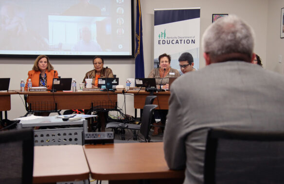 Three women sit around a table in a conference room listening to a man seated at a table in front of them.