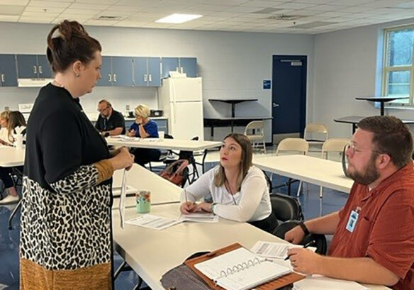 Two people sit on one side of a table while another person stands up on the other side