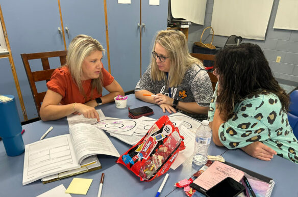 Three people talk while sitting at a desk with books, papers and a bag of candy on it.