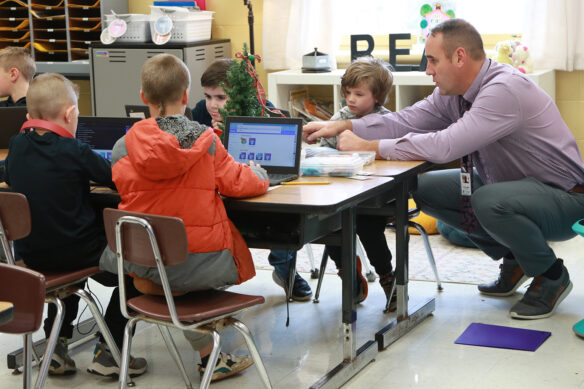 A man crouches down to help a group of students while they're working on computers