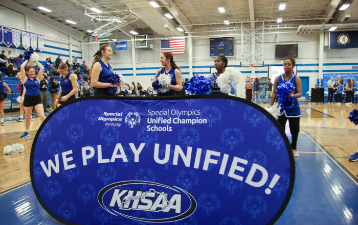 Cheerleaders stand behind a sign that says We Play Unified! in relation the Special Olympics Unified Champion Schools initiative