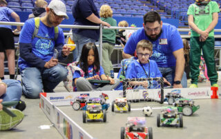 A couple of students, with adults watching over them, play a soccer game with robots