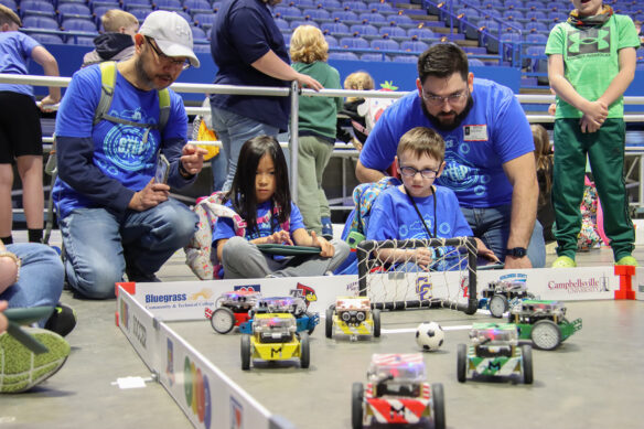 A couple of students, with adults watching over them, play a soccer game with robots