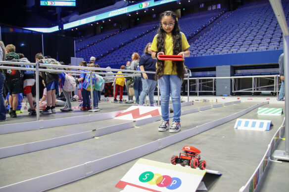 A girl holds a tablet that is controlling a robotic car that has a red hat with a M on it