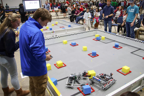 Students play with robots that are pushing colored blocks into boxes on the floor in a tic-tac-toe-like game