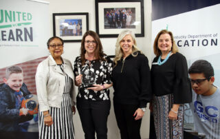 Four women stand in front of Kentucky Department of Education signs, with Emily Lehman holding a glass award.