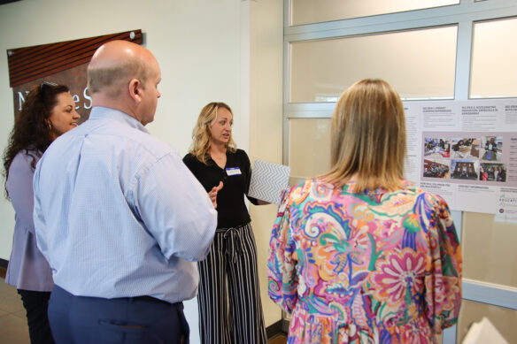 A woman talks to a group of people while looking at a poster with multiple pictures and words on it.