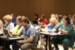 A group of educators are sitting at tables, writing in notepads