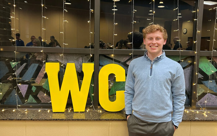 A person stands in front of a window with the letters "WC" behind him in gold.