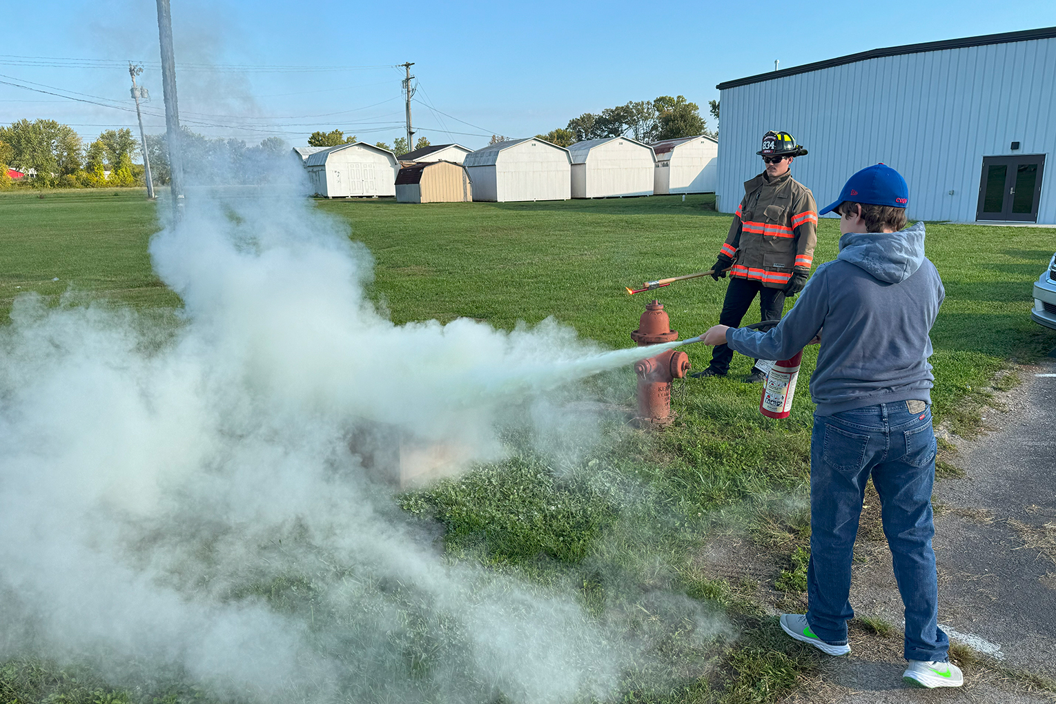 A student uses a fire extinguisher in a grassy field as a firefighter watches