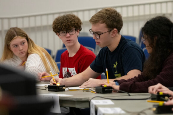 A group of students write with pencils on paper as two students look at one of the other students