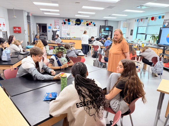 A teacher stands next to a table with students sitting around it in a classroom