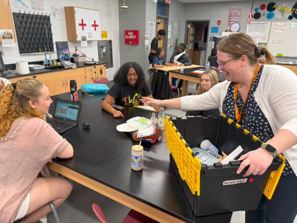 A smiling woman reaches for something on a table as she hovers over a box with recycling in it. Students are sitting around the table