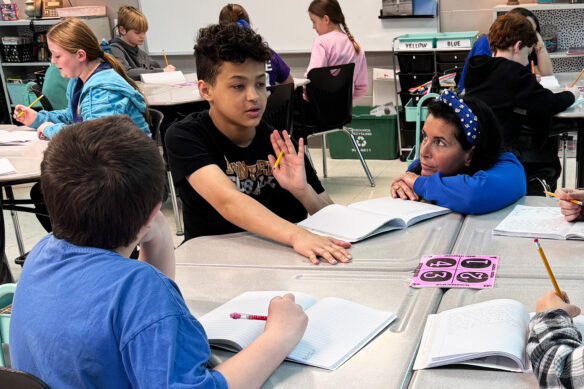 A woman crouches down by a table as multiple students work. One of the students appears to be speaking and is gesturing with his hand