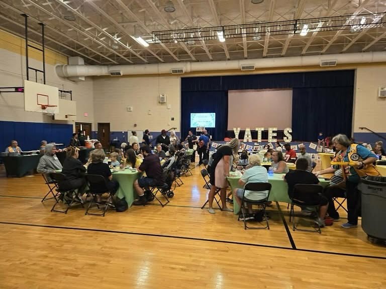 Groups of people sit at tables in a gymnasium with a display saying 'YATES' in the background