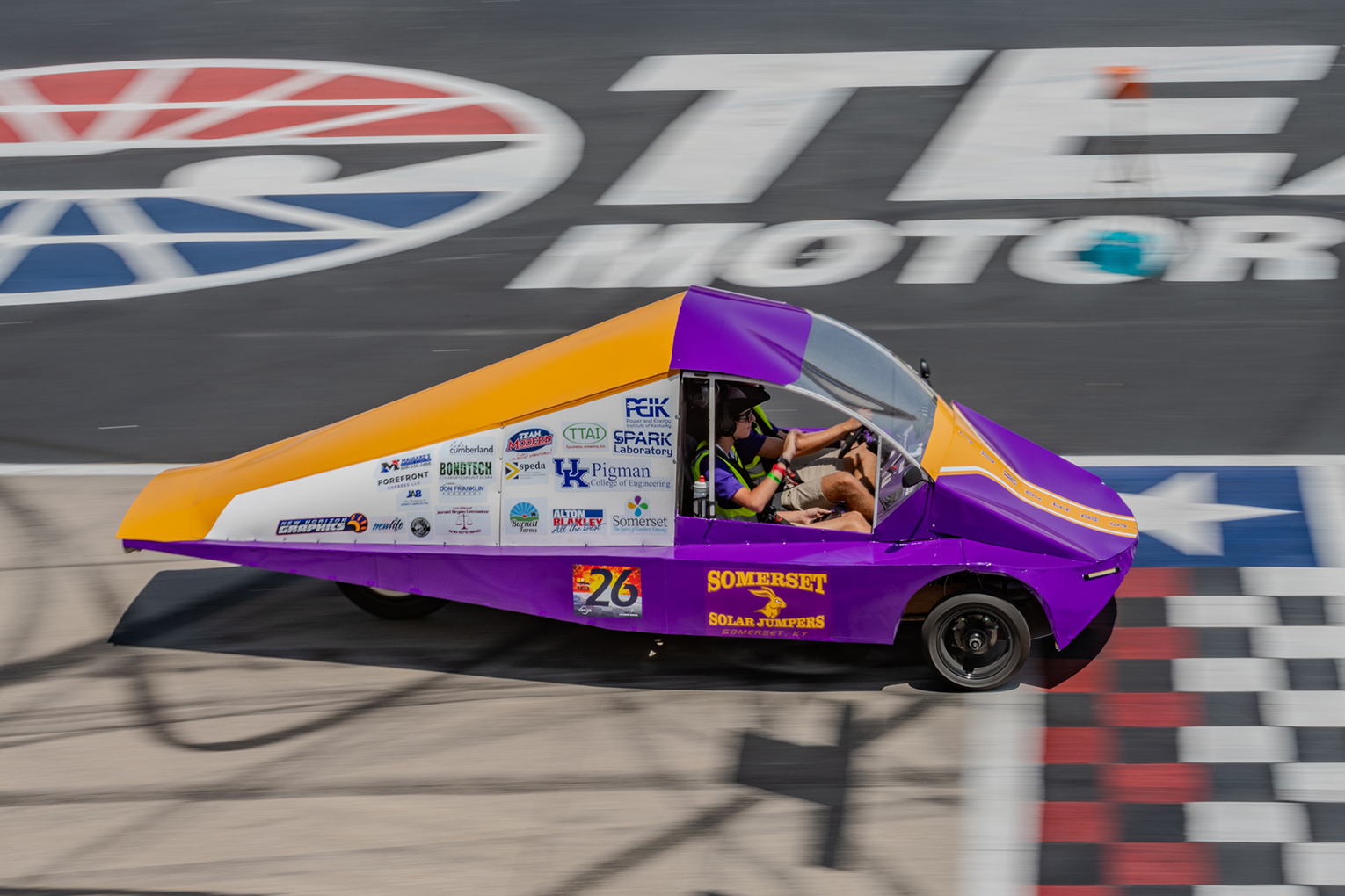 A solar-powered care with sponsorship decals on the side and two students inside crosses a finish line as the Texas Motor Speedway logo sits above them