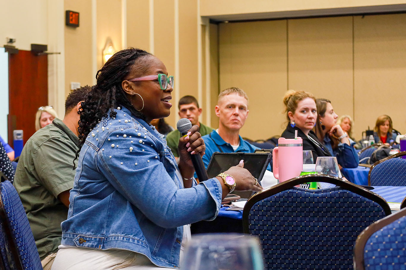 A woman holds a microphone in an audience to ask a question, while others seated at tables around her look on.