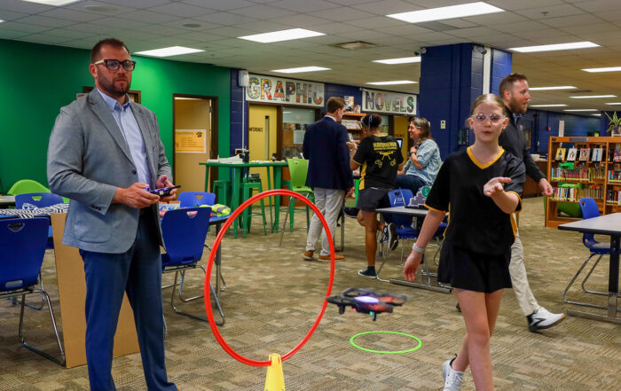 A man uses a remote controller to fly a drone through an obstacle course while a middle school student helps him