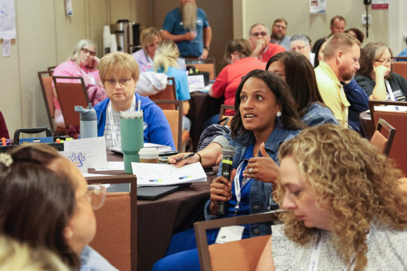 A woman speaks into a microphone while sitting in a room full of people sitting at tables
