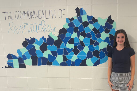 Harper Blake stands in front of a mural of Kentucky with each county painted a shad of blue with the words "The Commonwealth of Kentucky" written above it