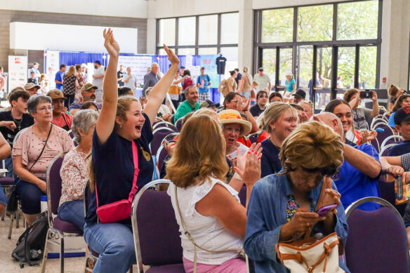 In a crowd of people sitting and clapping, one girl raises her hands and has a surprised expression on her face