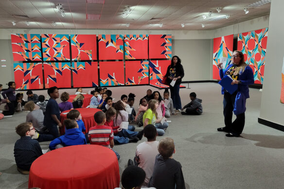 A woman speaks to a room full of children who are sitting on the floor in a room inside the National Quilt Museum