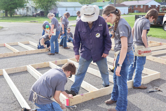 A kid kneels down and hammers a nail into a piece of wood as others look on