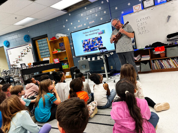 A man performs on a ukulele to a classroom of students who are sitting on the floor