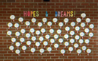 A brick wall features the words "Hopes & Dreams" with several papers cut into hot air balloon designs with writing on them and photos of kids