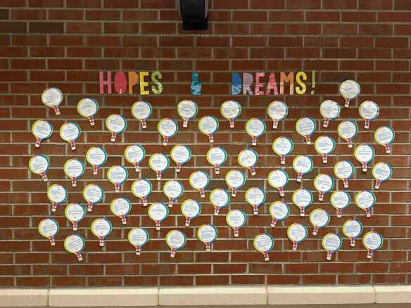 A brick wall features the words "Hopes & Dreams" with several papers cut into hot air balloon designs with writing on them and photos of kids