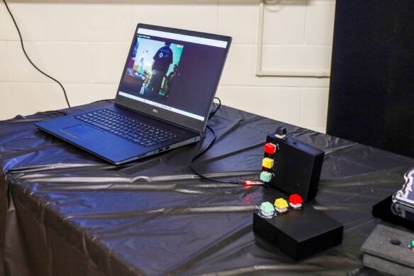 A laptop sits on a table with a video streaming a camera of the classroom. Two black boxes sit next to the laptop, both featuring red, yellow and green buttons on one side and a black button on another side