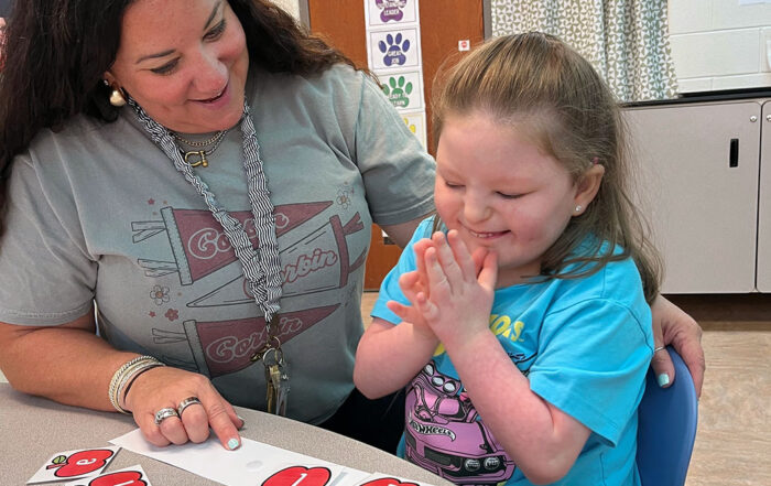 Melanie Howard smiles sitting next to a student, who is clapping and starting down at letters on a the table and clapping.