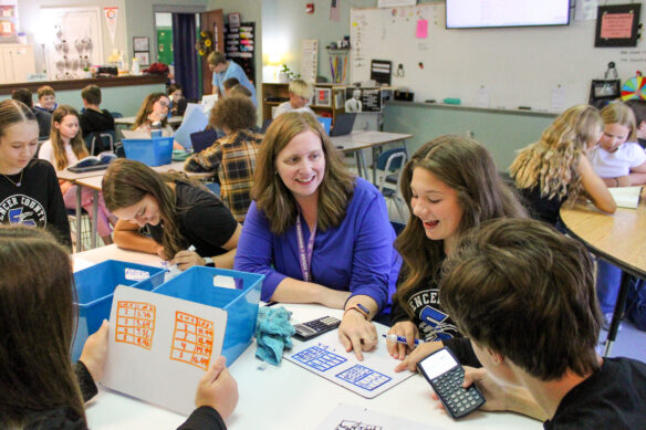 A smiling Michelle Gross sits at a table with students who are working on math problems.