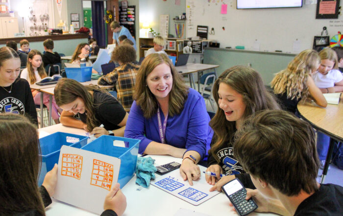 A smiling Michelle Gross sits at a table with students who are working on math problems.