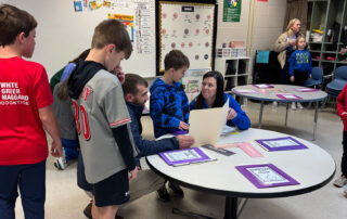 A woman sits at a table and shows something on a folded piece of paper to multiple students standing next to her