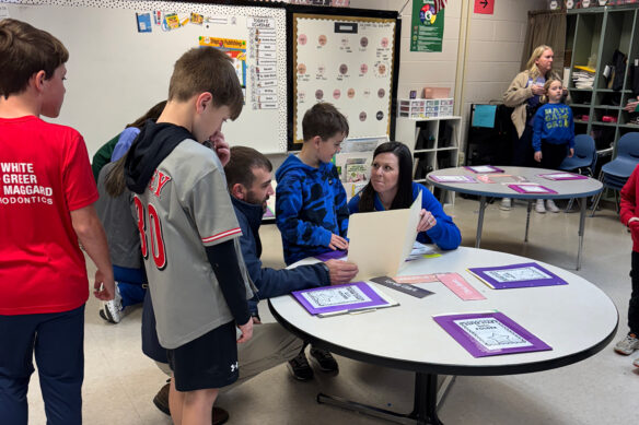 A woman sits at a table and shows something on a folded piece of paper to multiple students standing next to her