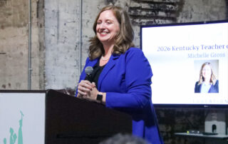 Michelle Gross stands at a podium, holding a microphone and smiling.