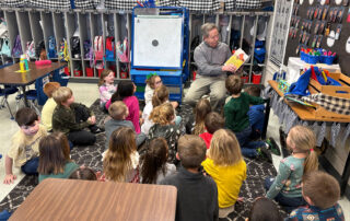 A man holds a book and reads to kids who are sitting on the floor around him in a classroom