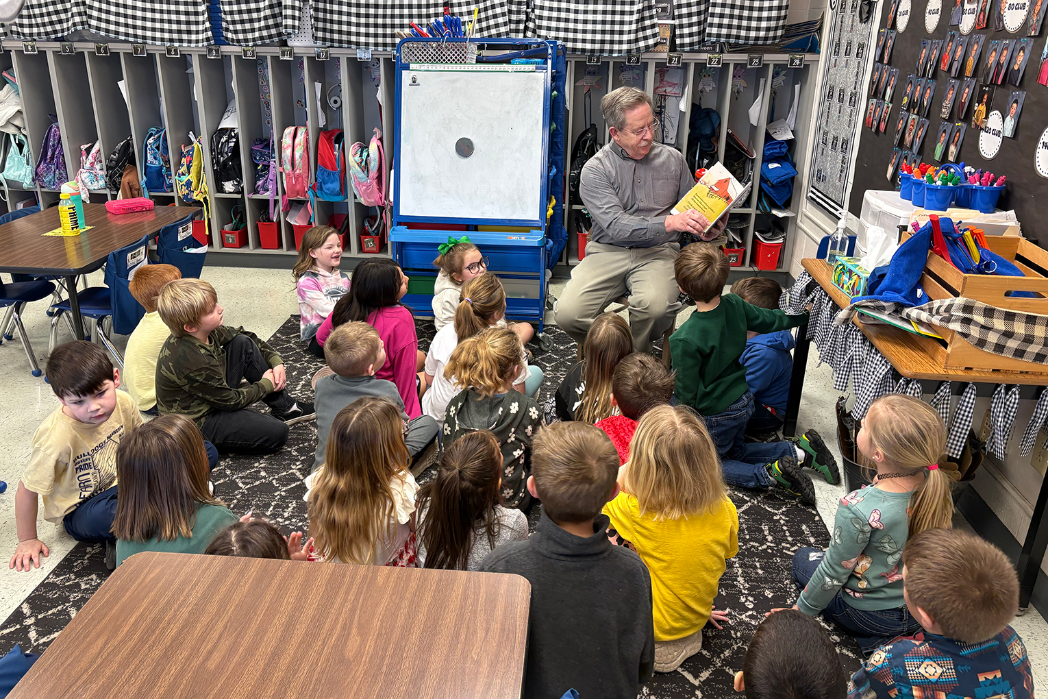 A man holds a book and reads to kids who are sitting on the floor around him in a classroom