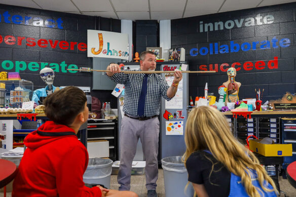 Scott Johnson holds pieces of wood in front of a classroom with kids watching him