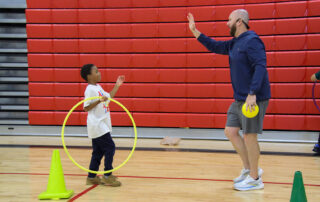 A teacher raises his hand for a high five with a student. The teacher is holding a yellow ball while the child is holding a yellow hula hoop. They're inside a gymnasium with red bleachers behind them, collapsed against a wall