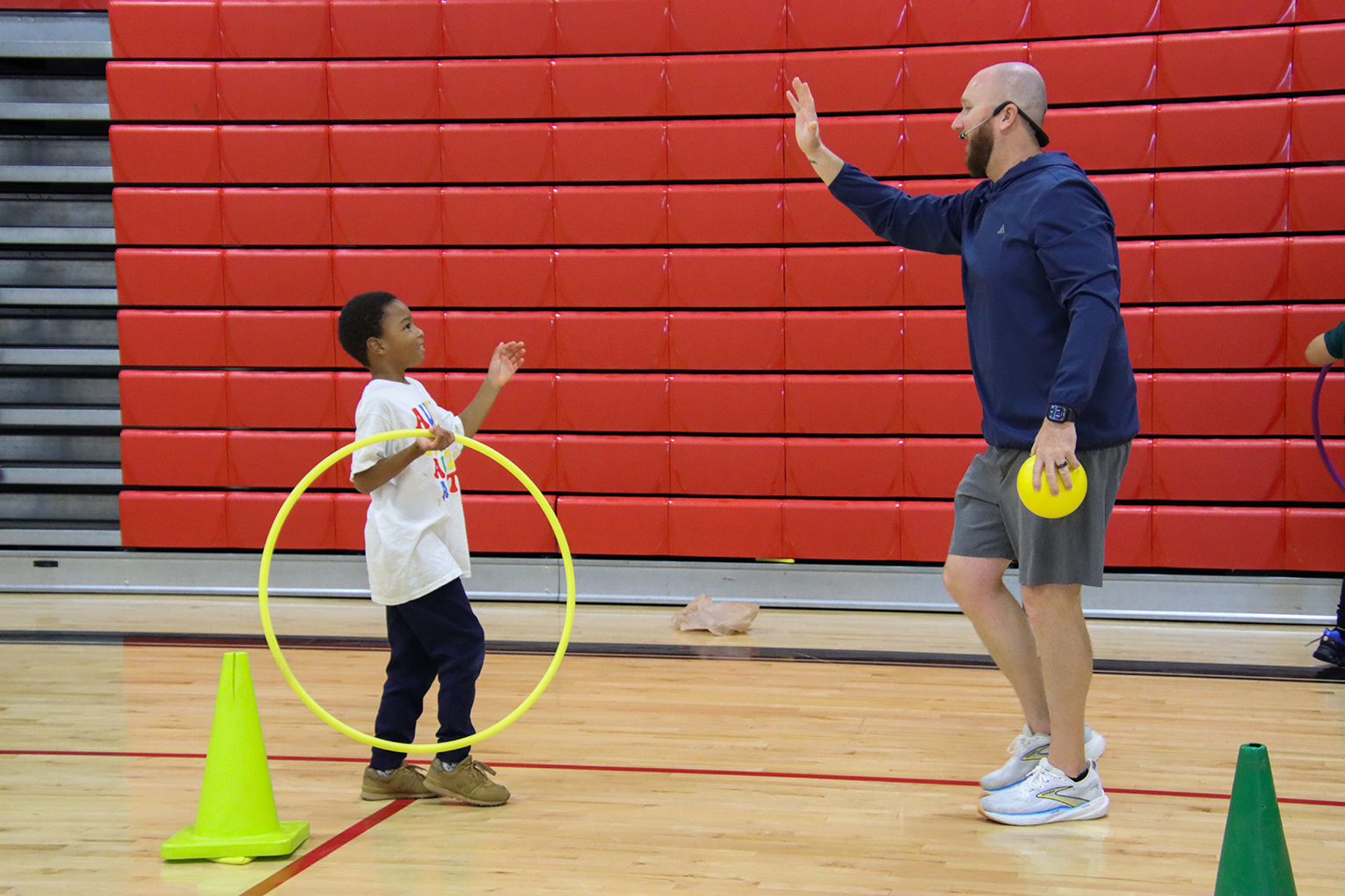A teacher raises his hand for a high five with a student. The teacher is holding a yellow ball while the child is holding a yellow hula hoop. They're inside a gymnasium with red bleachers behind them, collapsed against a wall