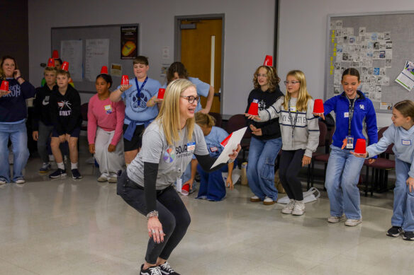 A woman crouches down while reading off a paper and holding her hand out above the ground as multiple kids in the background mimic her while holding red cups